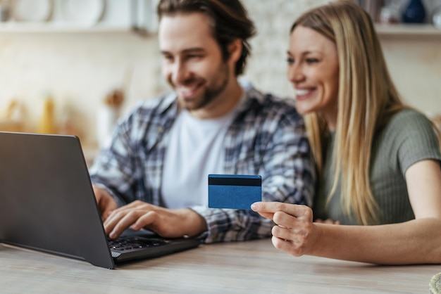 Glad young caucasian couple shopping online and show credit card in kitchen interior blurred