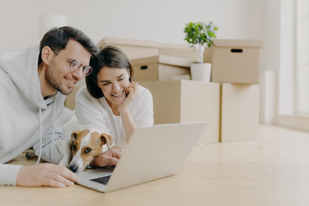 Glad young female and male discuss home repair projects look attentively into laptop their dog lies near pose in living room on floor dressed in casual clothes People and new home concept