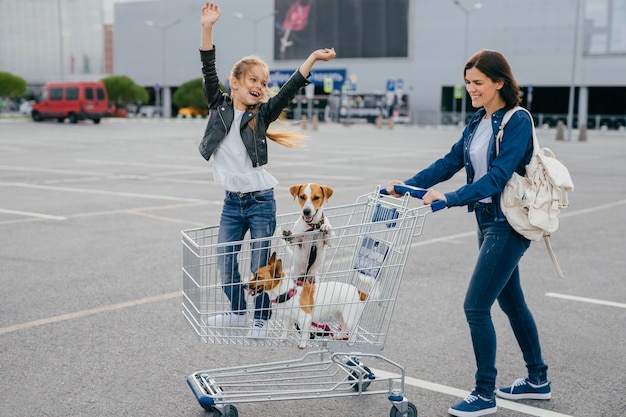 Glad young mother, daughter and their two dogs in shopping cart return home from mall