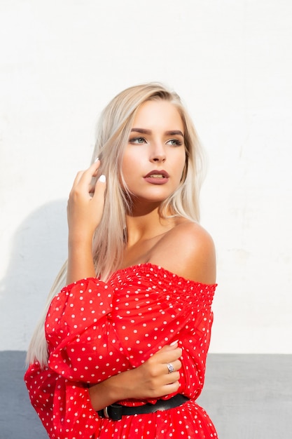 Glamor blonde model with naked shoulders in red dress, posing on the in soft sunset light