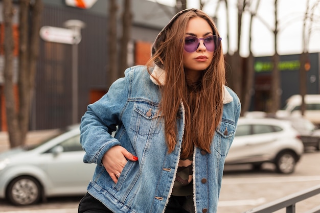 Glamorous beautiful young hipster woman in trendy purple glasses in oversized blue jeans jacket with long hair is resting near parking in the city. Modern urban girl model in stylish clothes outdoors.