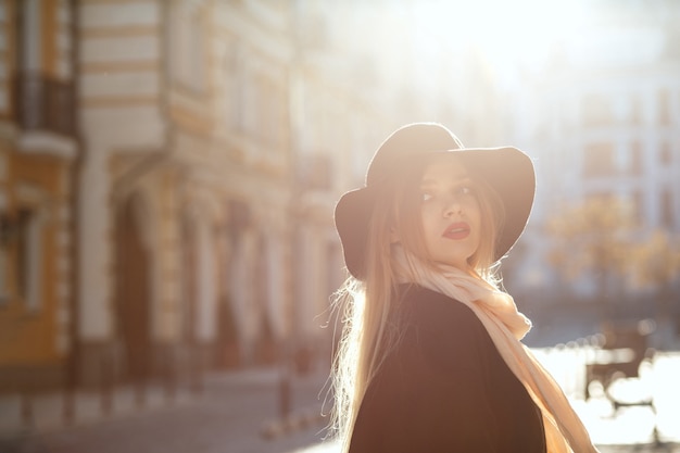 Glamorous blonde girl wearing hat, scarf and coat, walking down the old street in sun rays. Space for text