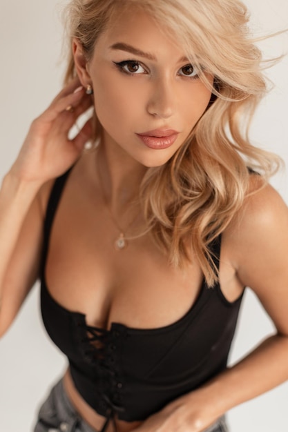 Glamorous portrait of a beautiful young girl with blond hair in a fashionable black sexy bra top in the studio on a white background looking at the camera