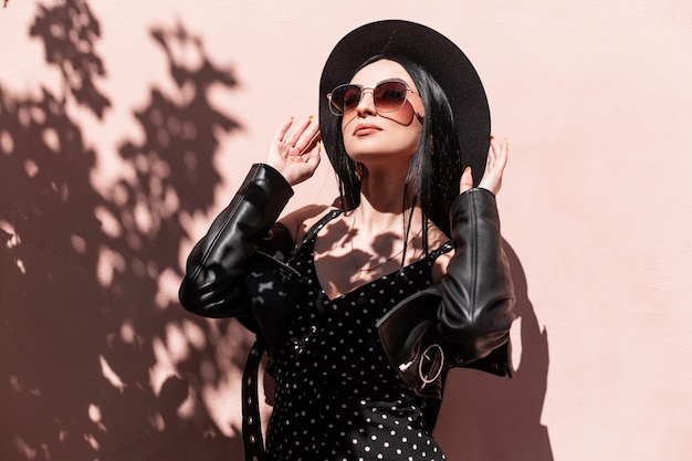 Glamorous young brunette woman in beautiful fashionable black clothes straightens hat outdoors. Luxurious sexy girl in dress in leather jacket in sunglasses posing and enjoys sunlight. Summer style.