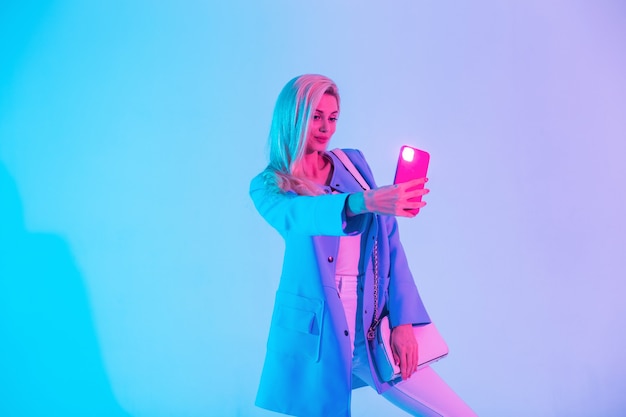 Glamorous young girl in fashionable bright clothes with a stylish handbag makes a selfie photo on a smartphone in the studio on a colored neon light