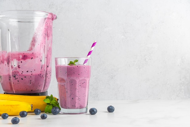 Glass of blueberry and banana smoothie with fresh juicy fruits and blender for making healthy summer