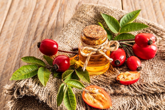 A glass bottle with a cork with natural rosehip seed oil on a burlap and a village table among freshly picked rose hips organic product