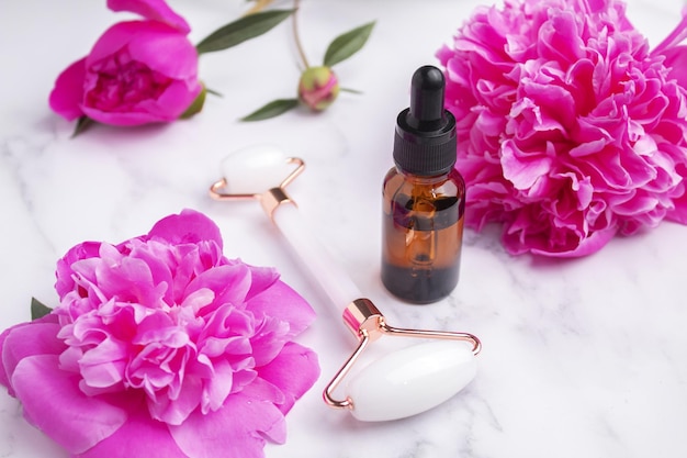 Glass bottle with oil roller for face massage on marble background with peony flowers