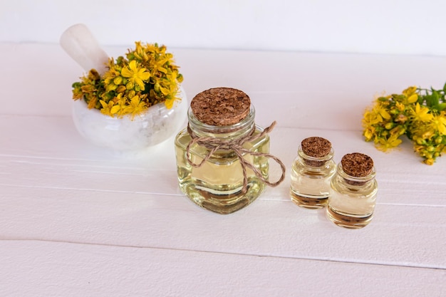 Glass bottles of organic St John's wort oil stand on a table of white wooden boards mortar and pestle with grass flowers alternative medicine