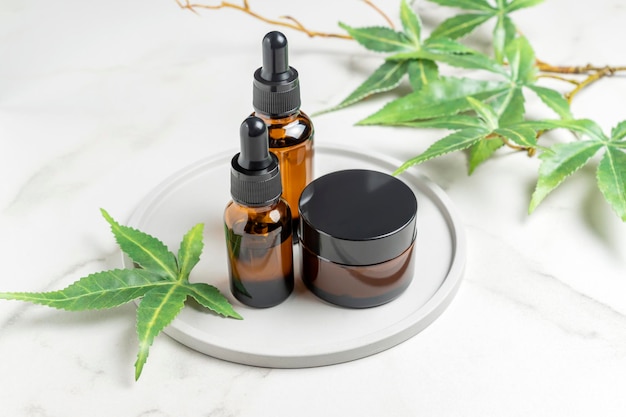 Glass bottles with CBD oil THC tincture and hemp leaves on tray a marble background Cosmetics cannabis oil and cream