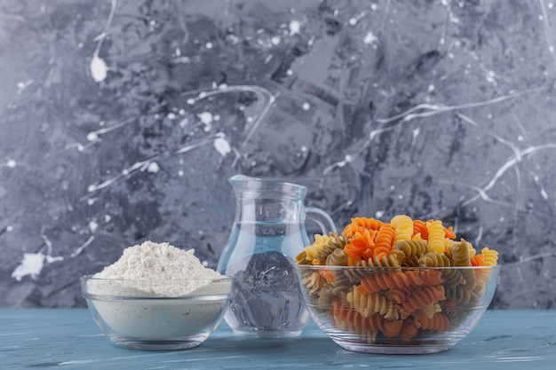 A glass bowl of multi colored raw spiral pasta with a pitcher and flour .