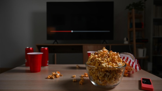 Glass bowl of popcorn and cups and remote control on wooden table Family vacation recreation relaxation and hobby concept