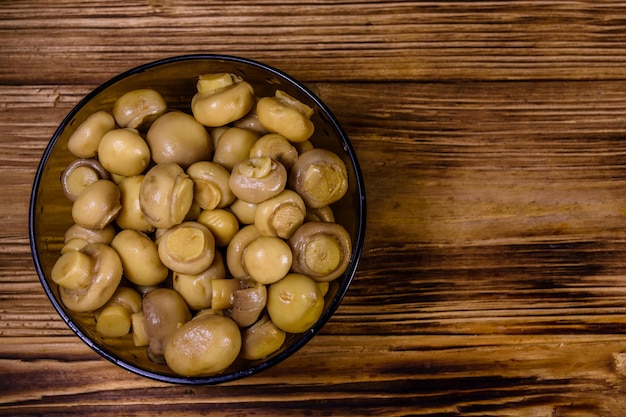 Glass bowl with canned mushrooms on rustic wooden table. Top view