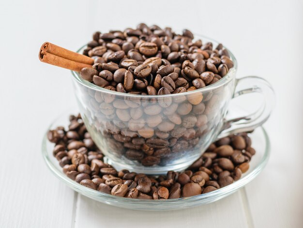 Glass bowl with coffee beans and cinnamon stick on white wooden table.