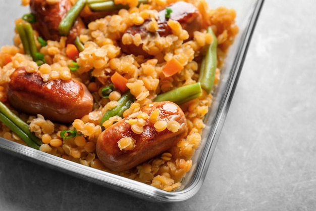 Glass bowl with tasty lentil porridge sausages and vegetables on kitchen table closeup