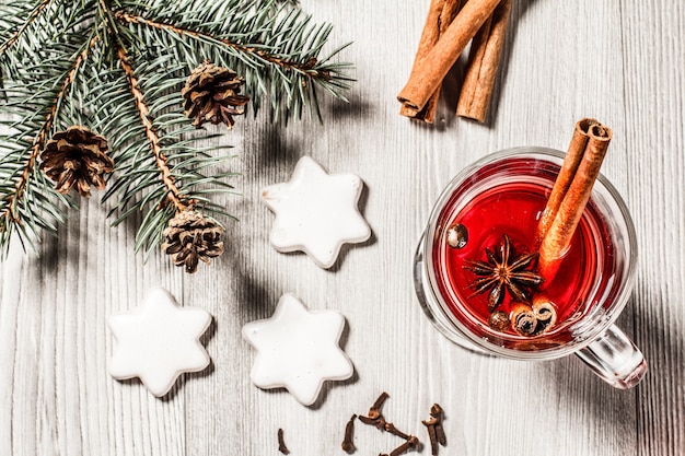 Glass of Christmas mulled wine with cinnamon, star anise and cloves on wooden background with white biscuits, natural fir tree branches and cones. Top view. Color toning effect.