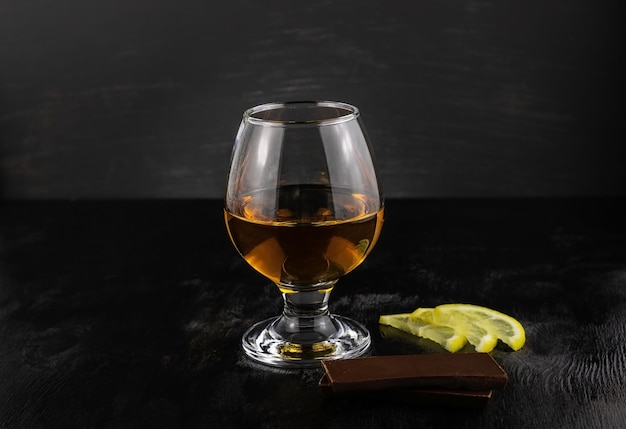 A glass of cognac, chocolate and lemon slices on a dark wooden background