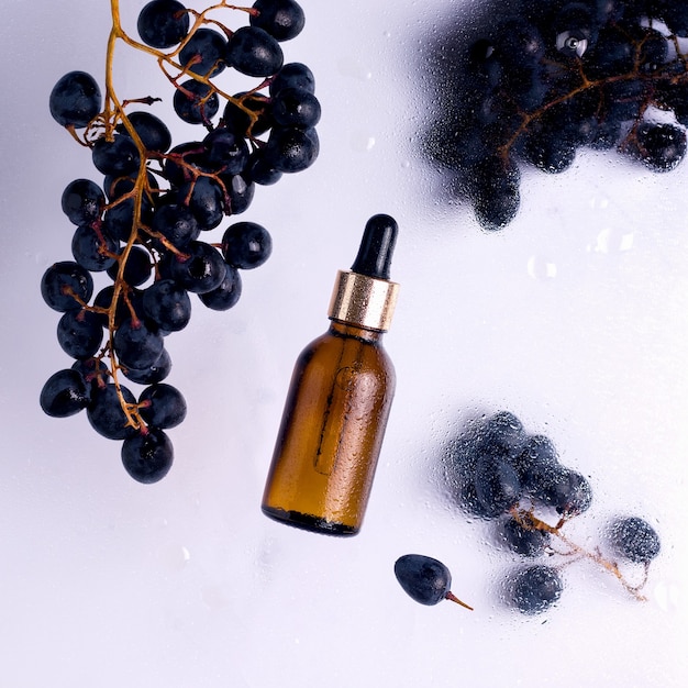 Glass cosmetic oils bottle with  black grapes around on wet trasnlucid background