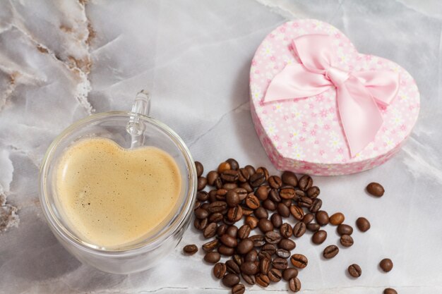 Glass cup of coffee and a gift box with roasted coffee beans on the kitchen table. The cup and the box in shape of a heart. Top view.