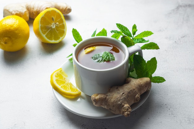 Glass cup of ginger tea with lemons and mint leaves on light surface,