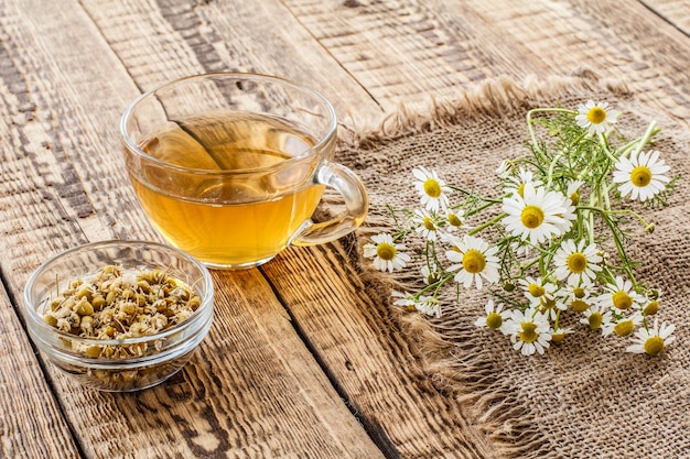 Glass cup of green tea, little glass bowl with dry flowers of matricaria chamomilla and fresh white chamomile flowers on wooden background.