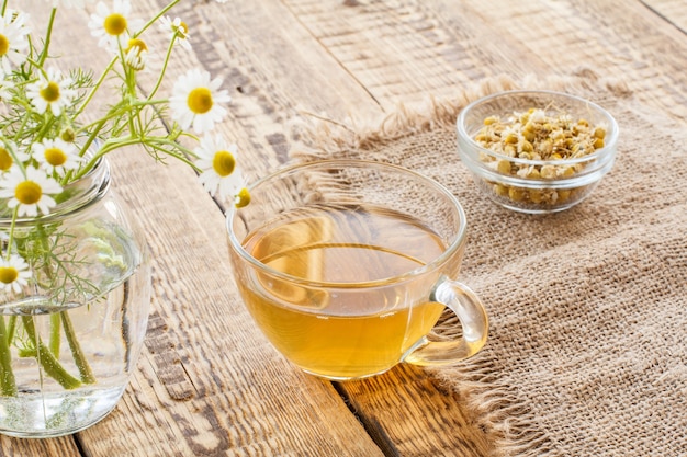 Glass cup of green tea, little glass bowl with dry flowers of matricaria chamomilla and fresh white chamomile flowers on wooden background.