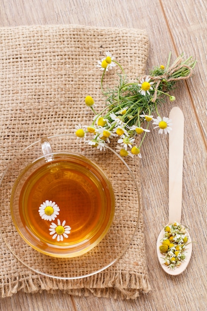 Glass cup of green tea with white chamomile flowers and sack on wooden background. Top view