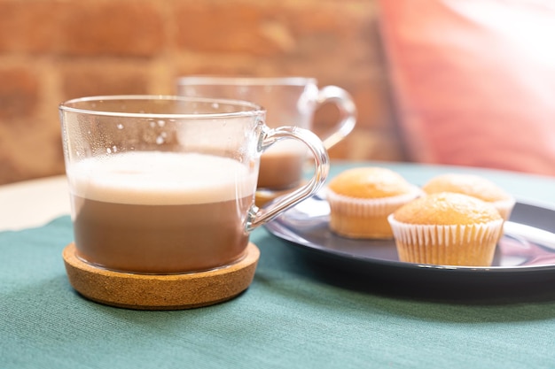 A glass cup of hot cocoa or cappuccino and three muffins on a plate standing on a table