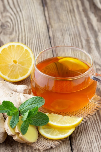 A glass cup of tea with lemon, mint and ginger on wooden rustic table.