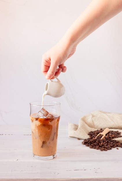 A glass cup with cold coffee and ice, coffee beans on a light surface. A hand pours cream and a glass