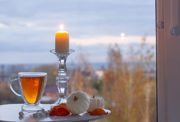 Glass cup with tea and burning candles on background window with autumnal landscape