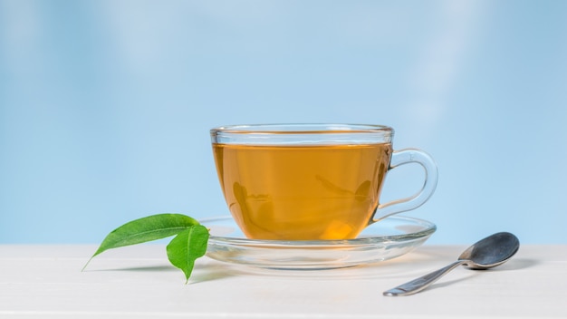 Glass Cup with tea and green leaves on a white table on a blue background. An invigorating drink useful for health.