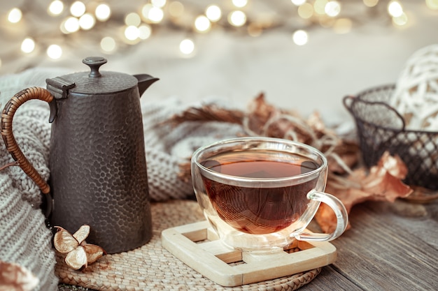 Glass cup with tea and teapot on wooden table with autumn decor details close up.