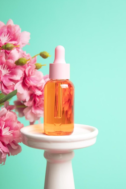 Glass dropper bottle on podium with flowers on light green background Cosmetic container mockup