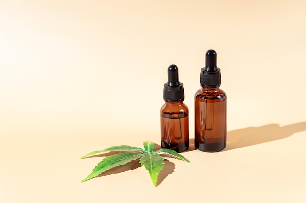 Glass dropper bottles with cbd oil on beige background with marijuana leaf Hemp based cosmetics Natural herbal hemp essential oil in a glass pipette bottle