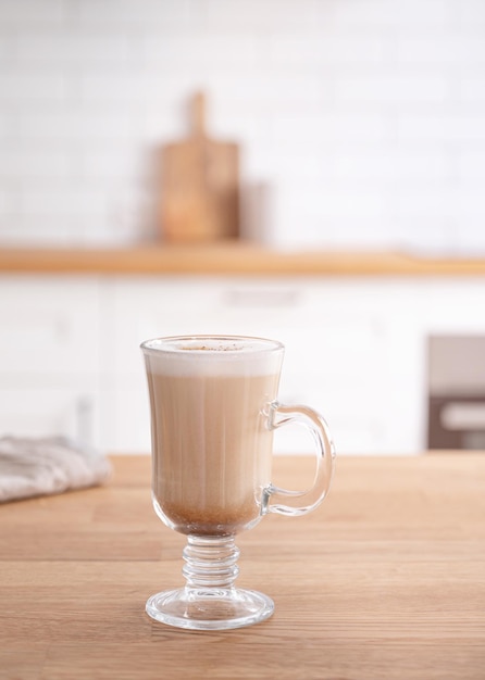 Glass of fragrant cappuccino coffee with cinnamon on a wooden table against the background of a white kitchen in the early morning Breakfast concept Front view