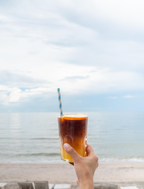 A glass of fresh iced orange americano decorated with sliced orange holding by hand on the beach view seascape vertical style Cold black coffee with orange juice mixing mocktail drink menu