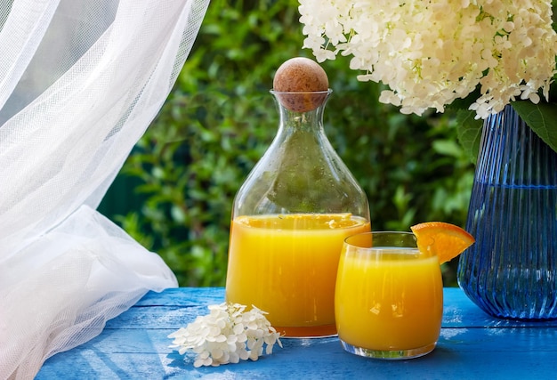 Glass of fresh orange juice garnished with a slice of ripe orange on the blue wooden table with flowers on background