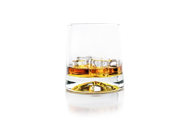 Glass of golden alcohol with ice cubes on white reflective surface, whiskey or cognac, isolated on white.