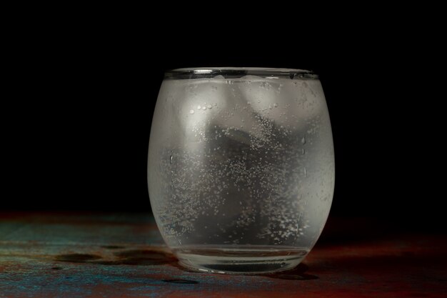 Glass of ice water filled with cold carbonated water on a dark background and rustic surface.