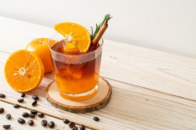 A glass of iced americano black coffee and layer of orange and lemon juice decorated with rosemary and cinnamon on wood background