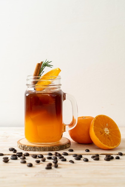 A glass of iced americano black coffee and layer of orange and lemon juice decorated with rosemary and cinnamon on wood background