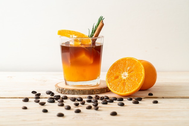 A glass of iced americano black coffee and layer of orange and lemon juice decorated with rosemary and cinnamon on wood table