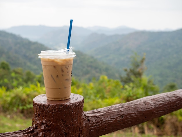 A glass of iced coffee is placed on the fence against a backdrop of mountains and sky