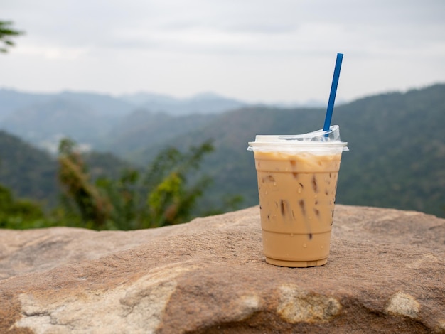 A glass of iced coffee is placed on a rock against a background of mountains and sky