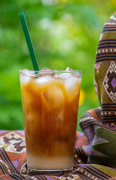 A glass of iced coffee with a green straw sits on a table with a teapot and a teapot in the background.