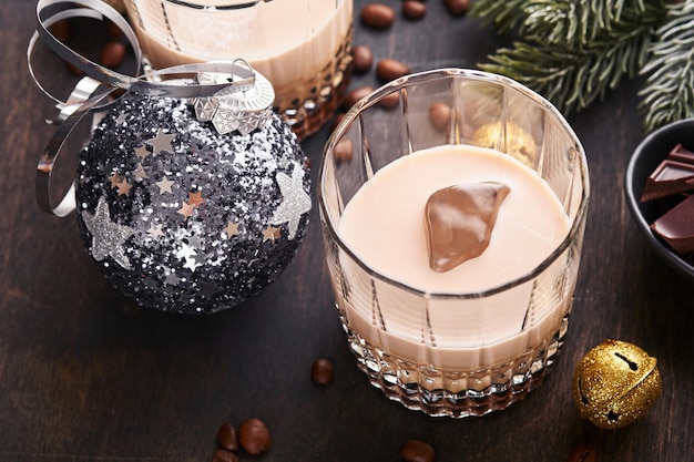 Glass of Irish cream baileys liqueur with ice, roasted coffee beans, cinnamon, Christmas decoration and chocolate on dark wood background table. Christmas table with gifts. Selective focus.