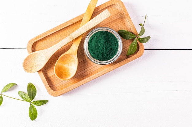 A glass jar filled with green spirulina algae powder and two wooden empty spoons on a wooden tray Top view White wooden table