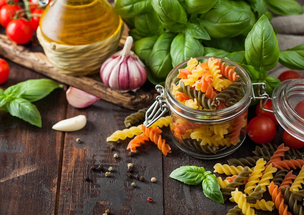 Glass jar of fresh raw tricolor fusilli pasta with oil and garlic basil plant and tomatoes on wooden background Top view