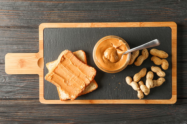 Glass jar with peanut butter and spoon, peanut, peanut butter sandwich and cutting board on wooden table, space for text and top view